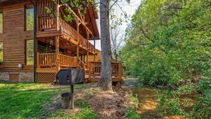 A wooden cabin with a balcony, a grill, and a stream in the background. - Creekside Dreams – Hot Tub, Fire Pit, Pool Table, 2 Bunk Beds & Creekside Deck (Pigeon Forge)