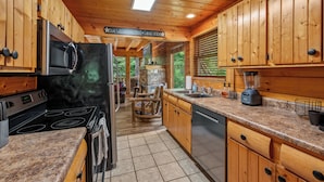 A kitchen with wooden cabinets, a granite countertop, a stove, a microwave, and a stone fireplace. - Creekside Dreams – Hot Tub, Fire Pit, Pool Table, 2 Bunk Beds & Creekside Deck (Pigeon Forge)