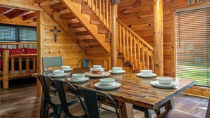 A wooden cabin interior with a dining table set for six, a staircase, and a cross on the wall. - Creekside Dreams – Hot Tub, Fire Pit, Pool Table, 2 Bunk Beds & Creekside Deck (Pigeon Forge)