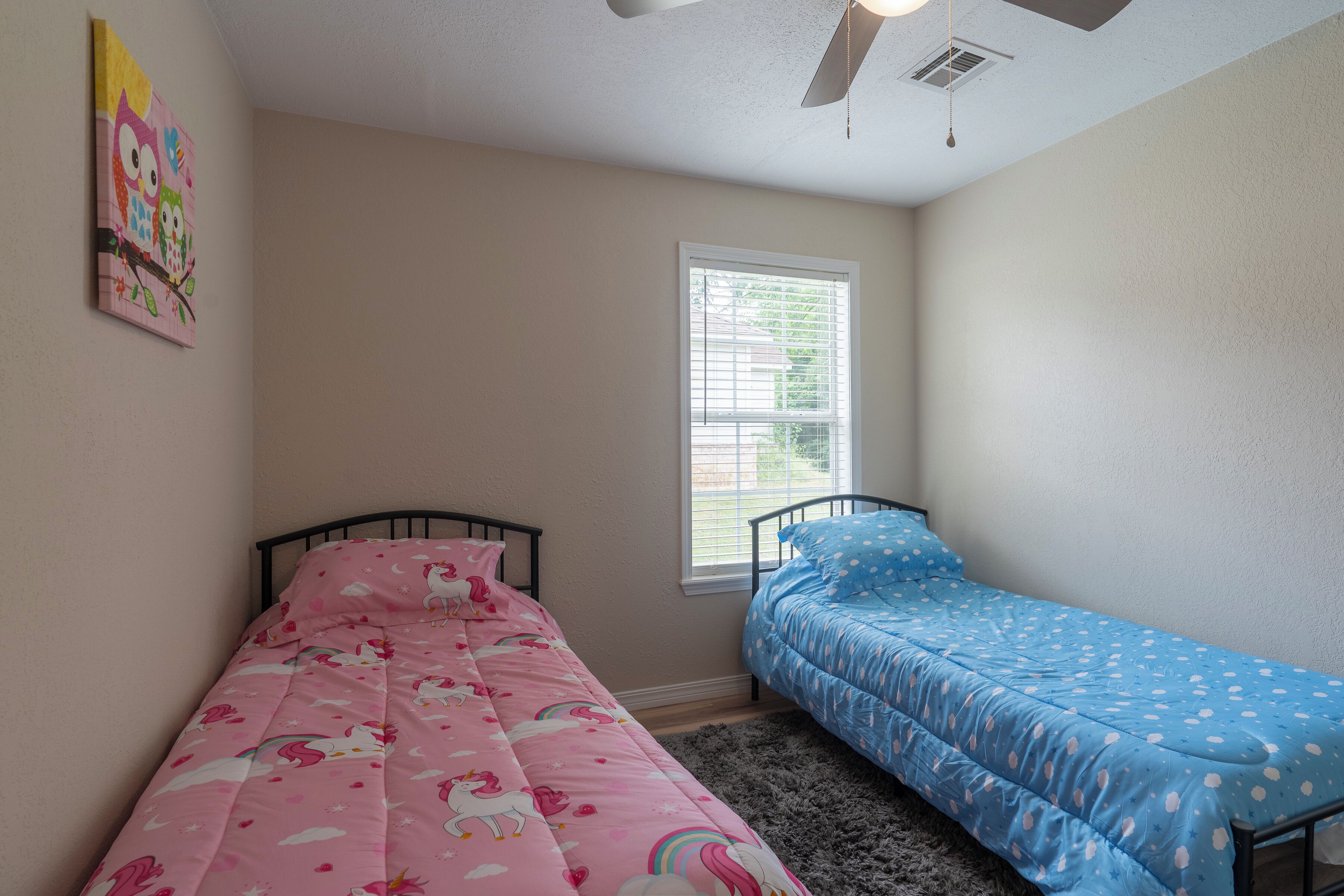 A bedroom with two beds, one pink and one blue, and a window with blinds.