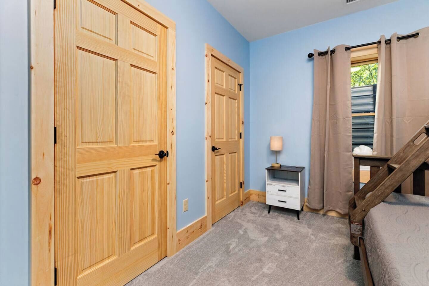 A bedroom with two wooden doors, a small bedside table with a lamp, a bed, and a window with curtains.