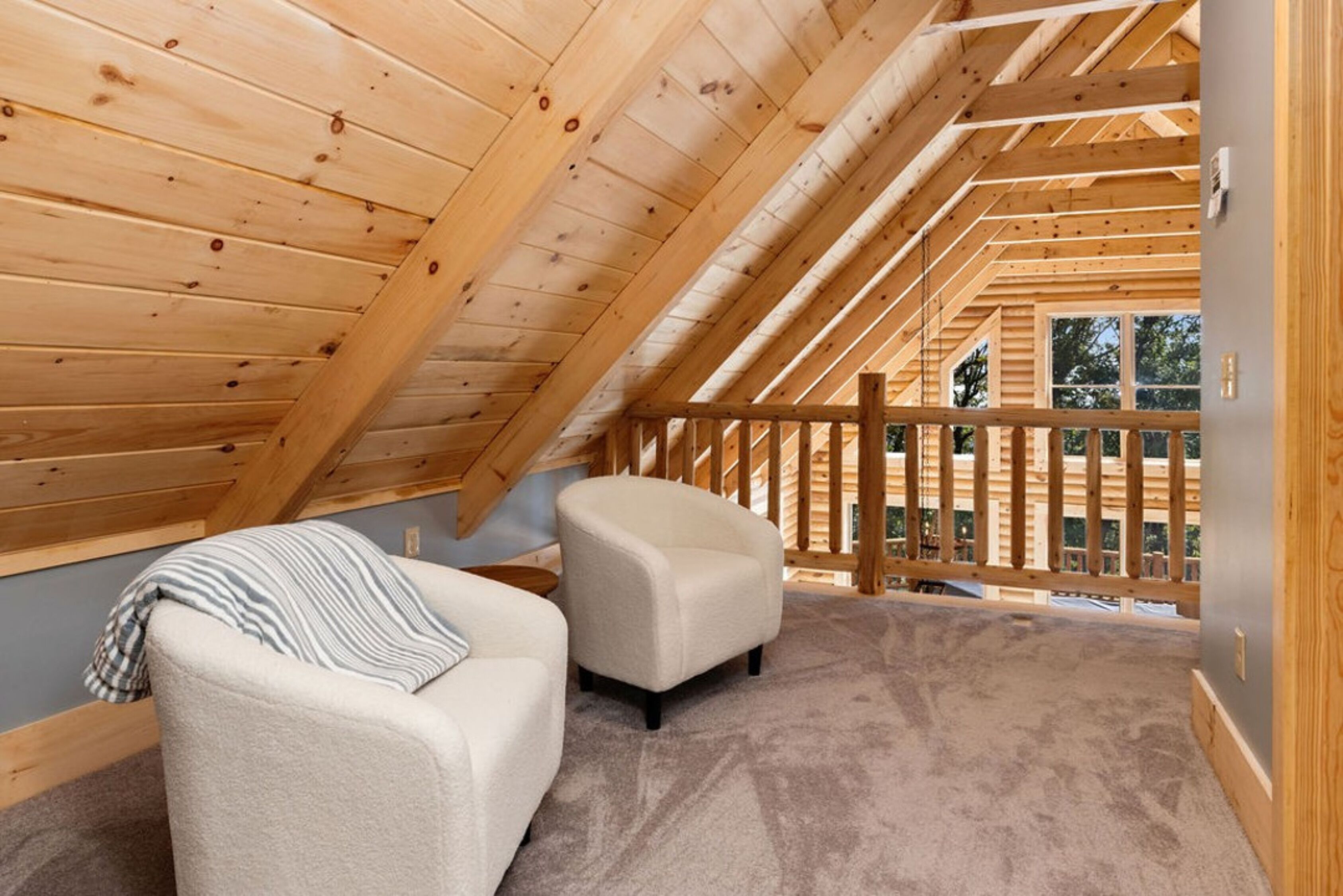 A cozy attic bedroom with a sloped ceiling, two plush armchairs, and a view of the outdoors through a wooden railing.
