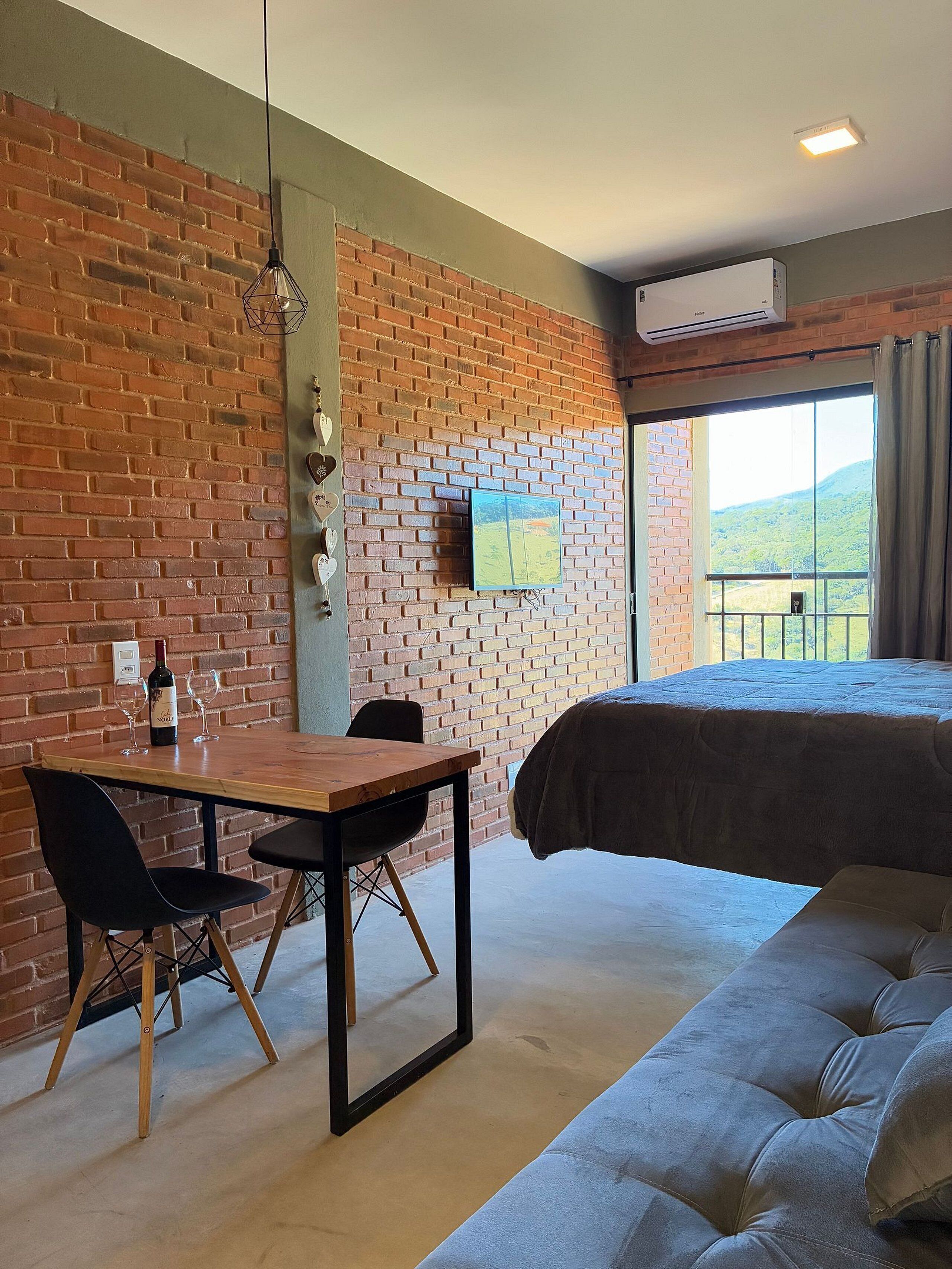 A room with a brick wall, a bed, a table with chairs, a TV, and a window with a view.