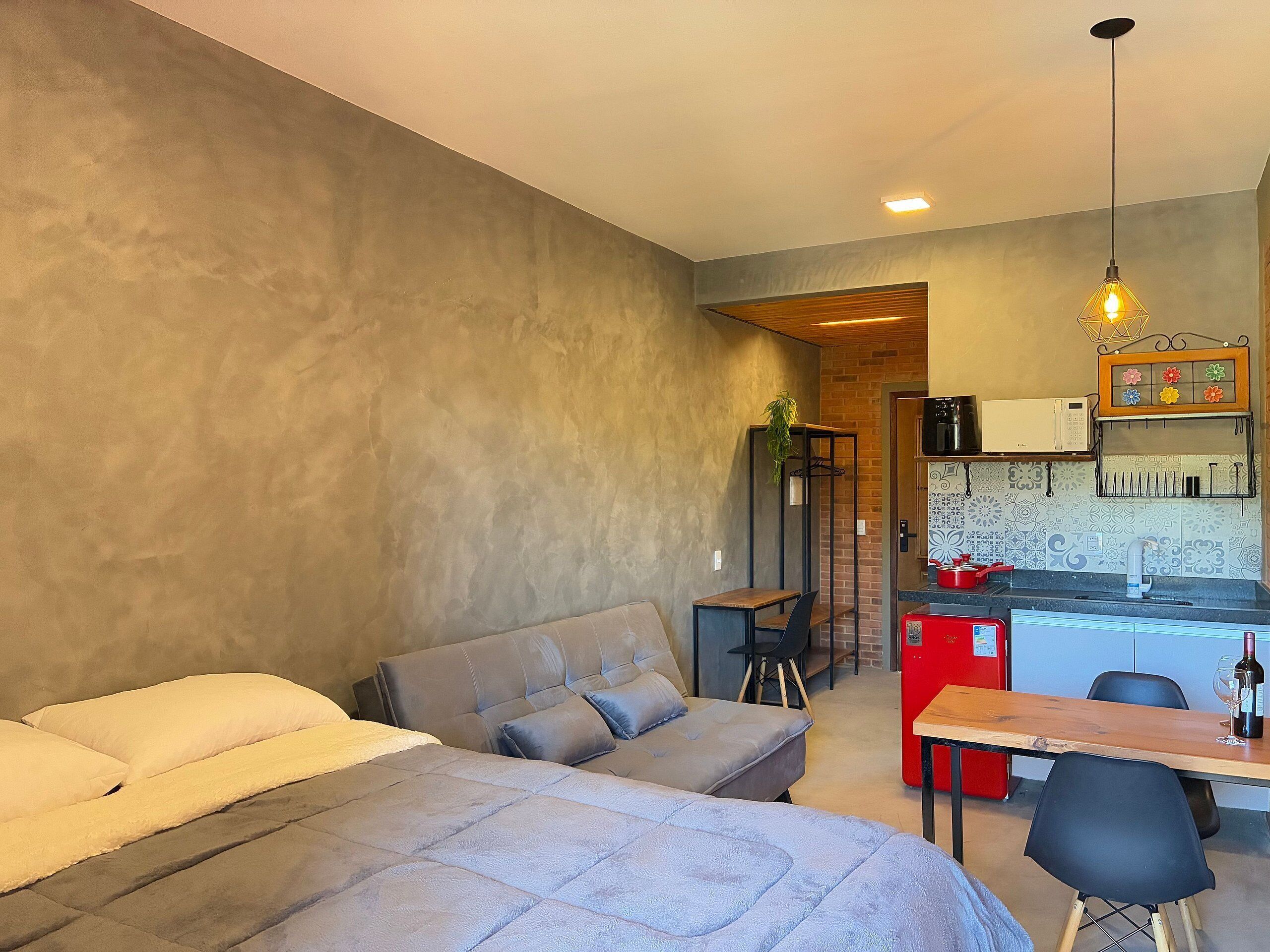 Quarto moderno com sofá-cama, mesa de madeira e cozinha compacta com geladeira vermelha.
