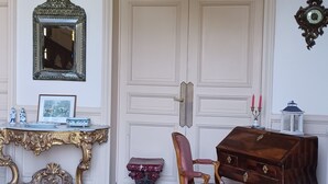 Ein Zimmer mit einer großen weißen Tür, einer Kristallleuchtre, einem Holztisch, einem roten Stuhl, einem kleinen Tisch mit einem roten Hocker und einem Spiegel an der Wand.