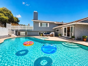Ein Schwimmbecken mit zwei aufblasbaren Ringen, eine Terrasse mit Gartenmöbeln und ein zweistöckiges Haus mit weißem Zaun.