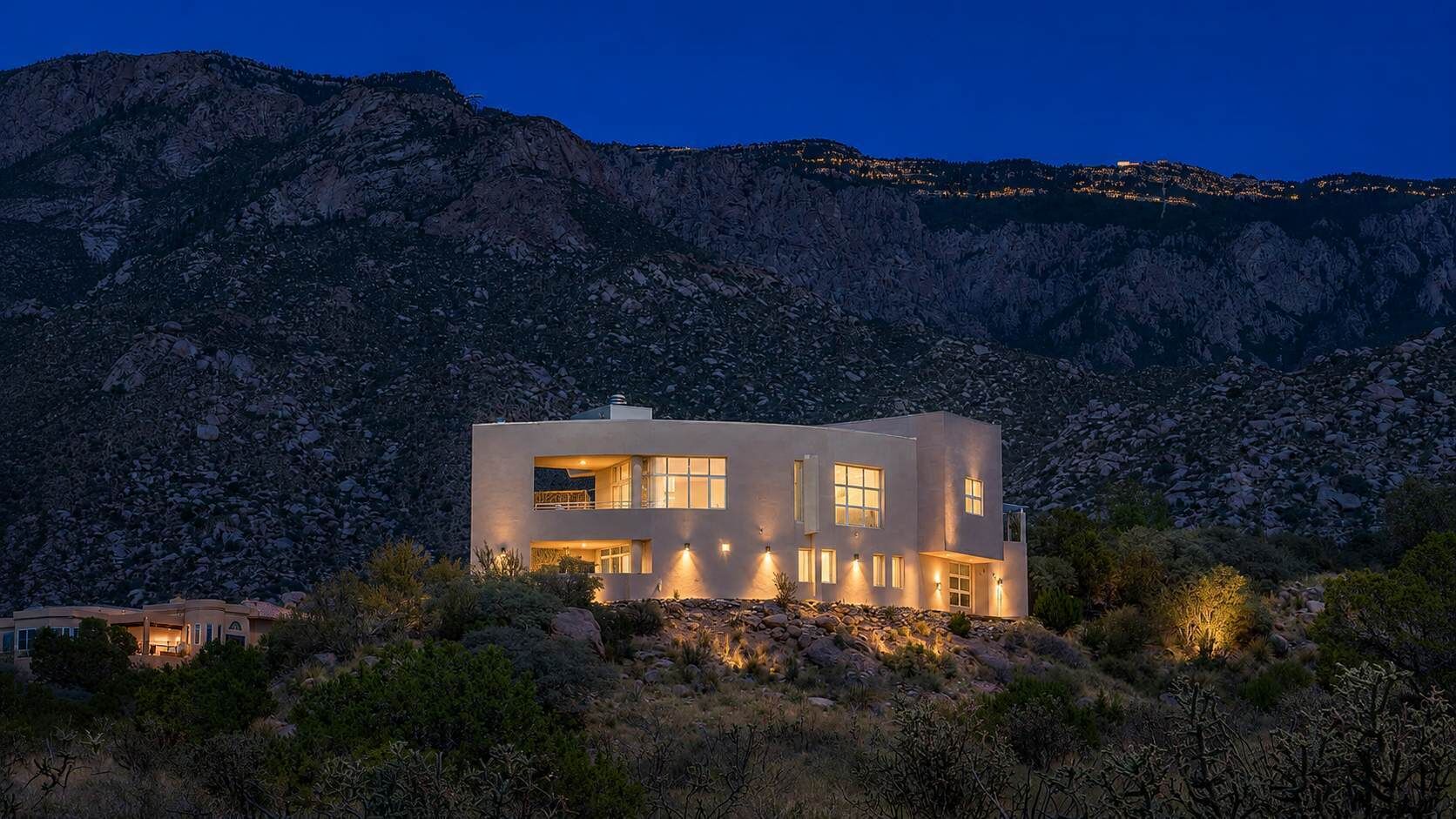 A modern two-story house with a flat roof, large windows, and a balcony, surrounded by rocky terrain and sparse vegetation.