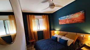A hotel room with a bed, a chair, a ceiling fan, a window with curtains, and a painting on the wall.