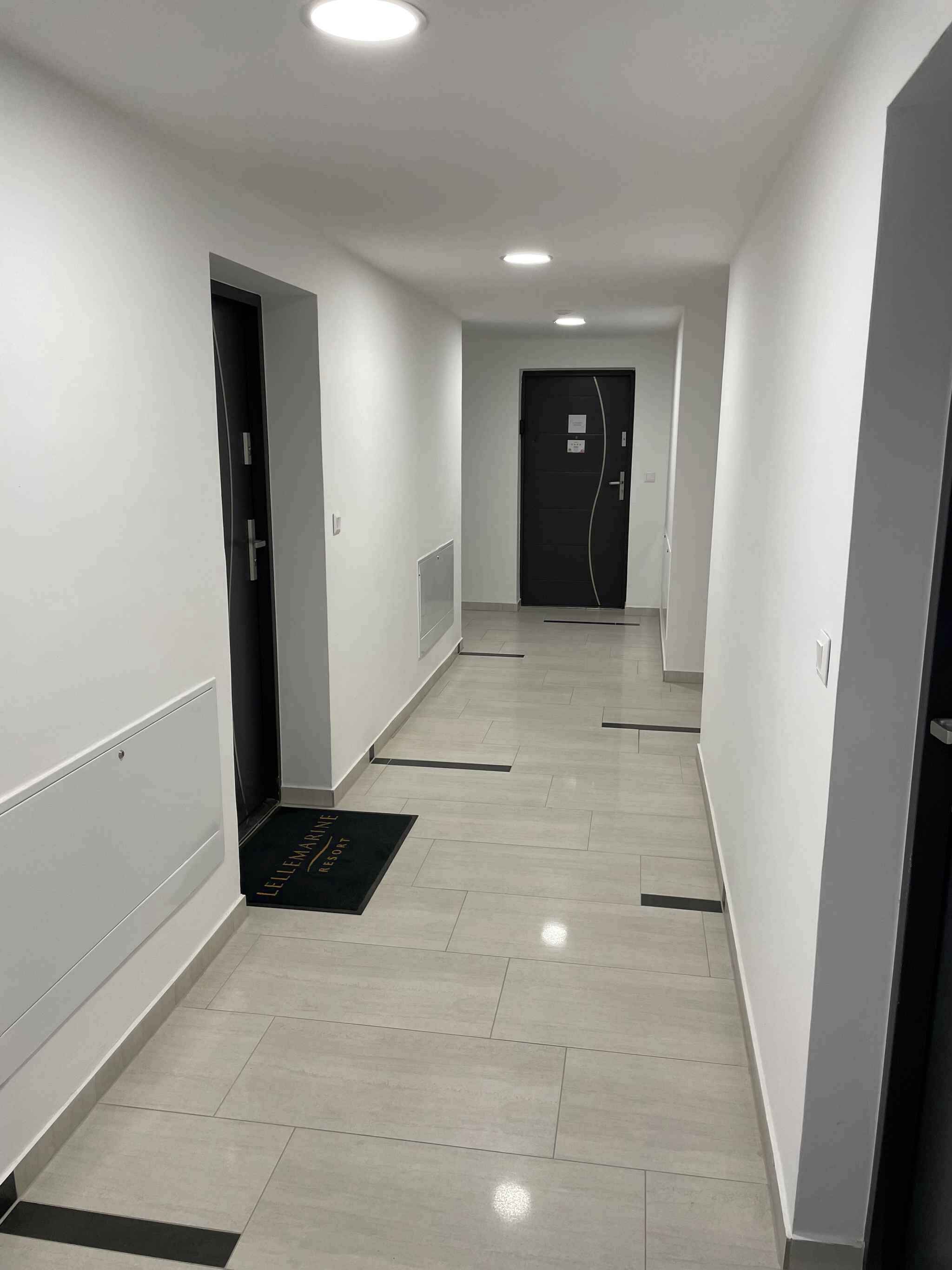 A hotel corridor with white walls, a tiled floor, and doors leading to rooms.