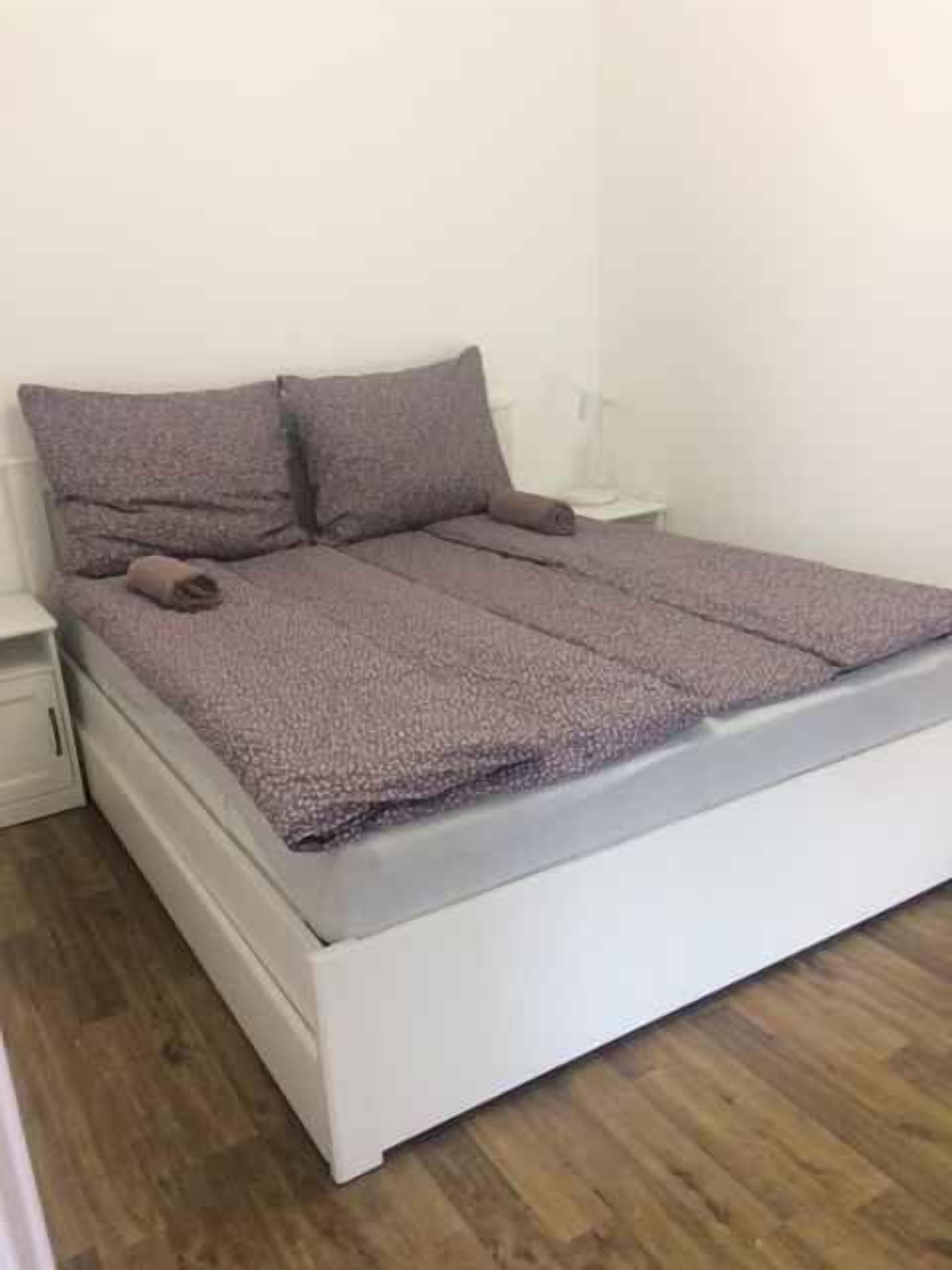 Een eenpersoonskamer met een wit bedframe, een houten vloer en een effen witte muur.