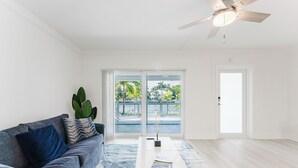 A spacious living room with a large window, a ceiling fan, and a view of a pool and palm trees. - Waterfront Retreat—Views, Pool, Hot Tub, Gym Walk to Beach & Boca Resort! (Boca Raton)