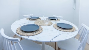 Una mesa redonda con cubierta de mármol blanco, rodeada de sillas blancas, con platos oscuros, individuales tejidos y cristalería transparente.