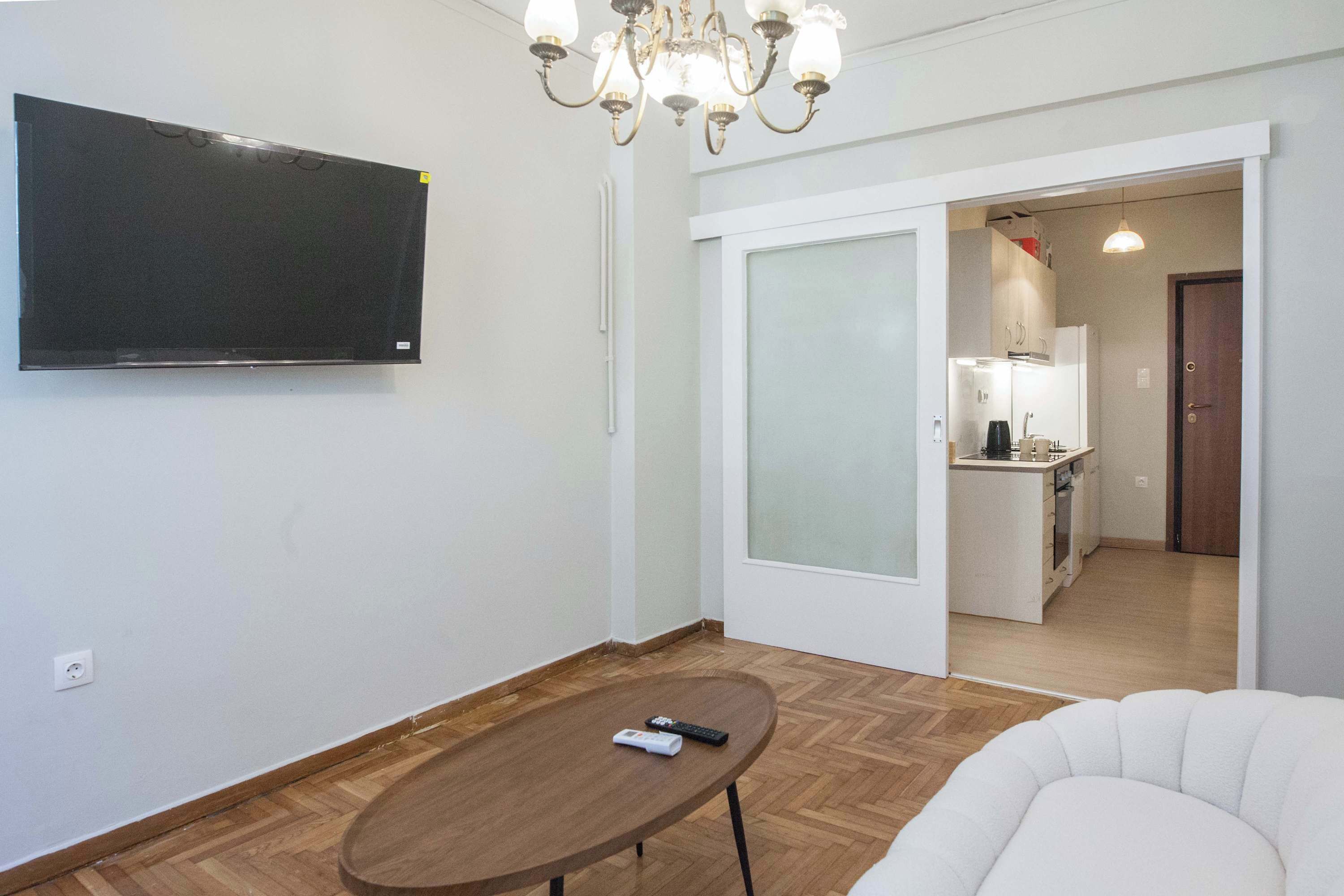 Ein modernes Wohnzimmer mit einem an der Wand befestigten Flachbildfernseher, einem Holztisch und einem weißen Sessel. Im Hintergrund ist eine Küche durch eine geöffnete Tür erkennbar.