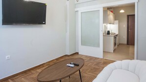 Una sala moderna con un televisor de pantalla plana montado en la pared, una mesa de centro de madera y un sillón blanco. Se ve una cocina a través de una puerta abierta al fondo.