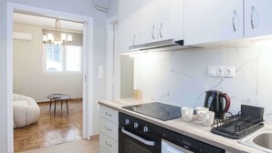 Una cocina moderna con armarios blancos, un horno negro y un lavavajillas. Hay una ventana con vistas al exterior, una araña y una pequeña mesa redonda con una silla.