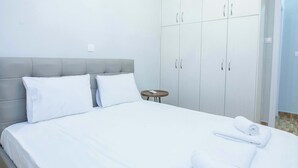 Una cama bien hecha con sábanas blancas, un cabecero tapizado en gris y una pequeña mesa redonda con un control remoto.