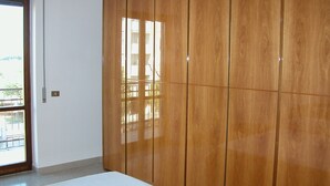 A room with a large wooden wardrobe, a door leading to an outdoor balcony, and a white textured bed.