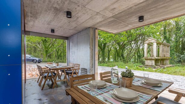 Un espace extérieur aménagé pour les repas, avec des tables et des chaises en bois, une vue sur un jardin et une statue, et une voiture garée à l’extérieur.
