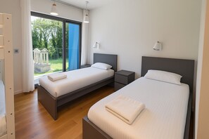 A modern bedroom with two beds, a small bedside table, a nightstand, and a large window with a view of greenery.