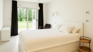 Une chambre moderne avec un grand lit, une petite table de chevet, une chaise, une table d’appoint en osier et une vue sur l’extérieur.