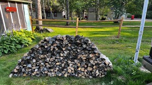 A pile of cut logs in a grassy area with a wooden fence and a shed in the background. - Charming 2-bedroom cabin on lake near Duluth MN with Air Conditioning (Saginaw)