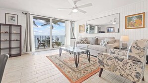 A spacious living room with a large glass coffee table, a sofa, a patterned armchair, and a ceiling fan.