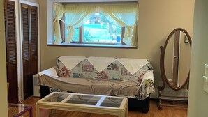 A cozy living room with a sofa, a glass coffee table, a window with curtains, a mirror, and wooden flooring.