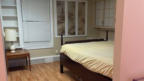 A bedroom with a bed, a nightstand, a lamp, a window with blinds, and a wall with a mural.