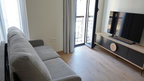 Una sala moderna con un sofá gris, un televisor de pantalla plana sobre una consola de madera y un ventanal corredizo que da a un balcón.