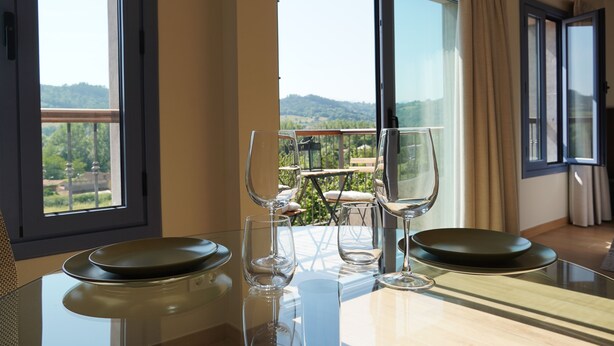 Un comedor con una mesa dispuesta para dos personas, vistas al exterior a través de amplias ventanas y un balcón con mobiliario.