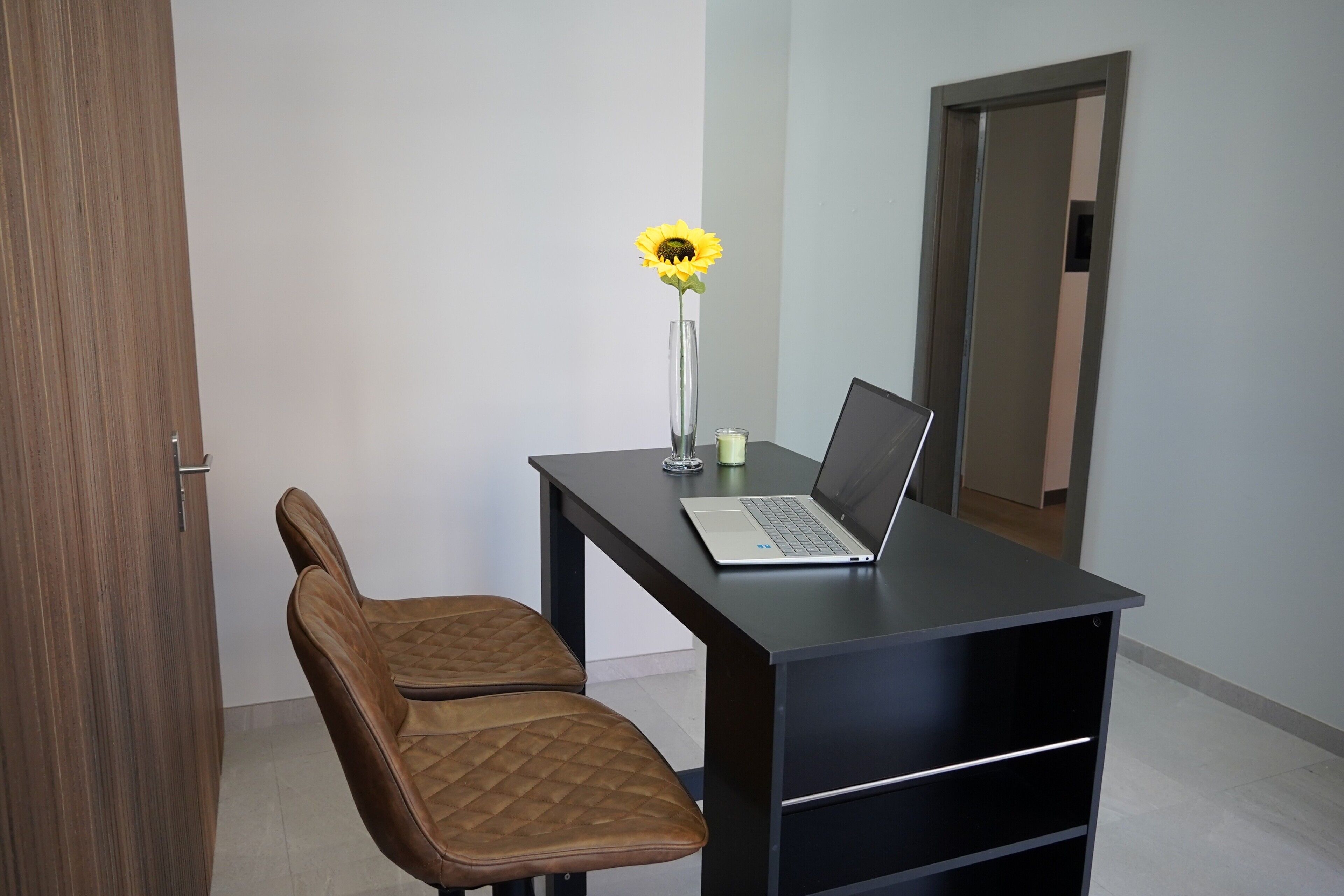 A room with a desk, a laptop, a chair, and a vase with a sunflower.