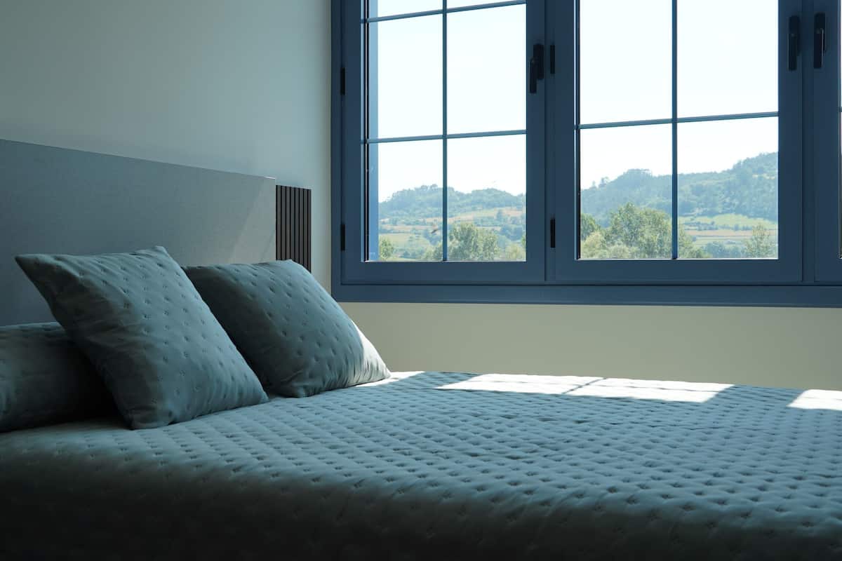 Un dormitorio con un ventanal grande, una cama con cubrecama acolchado y dos almohadas.