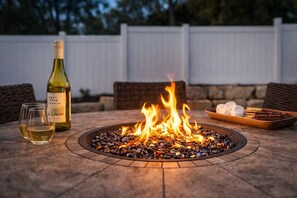 A fire pit with stones, a bottle of wine, glasses, and a tray of snacks on a patio. - NineSixFive Fieldhouse | Stadium · Arcades · Luxe (Ashwaubenon)