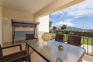 A balcony with a glass table, wicker chairs, a wicker lamp, and a view of a distant landscape.