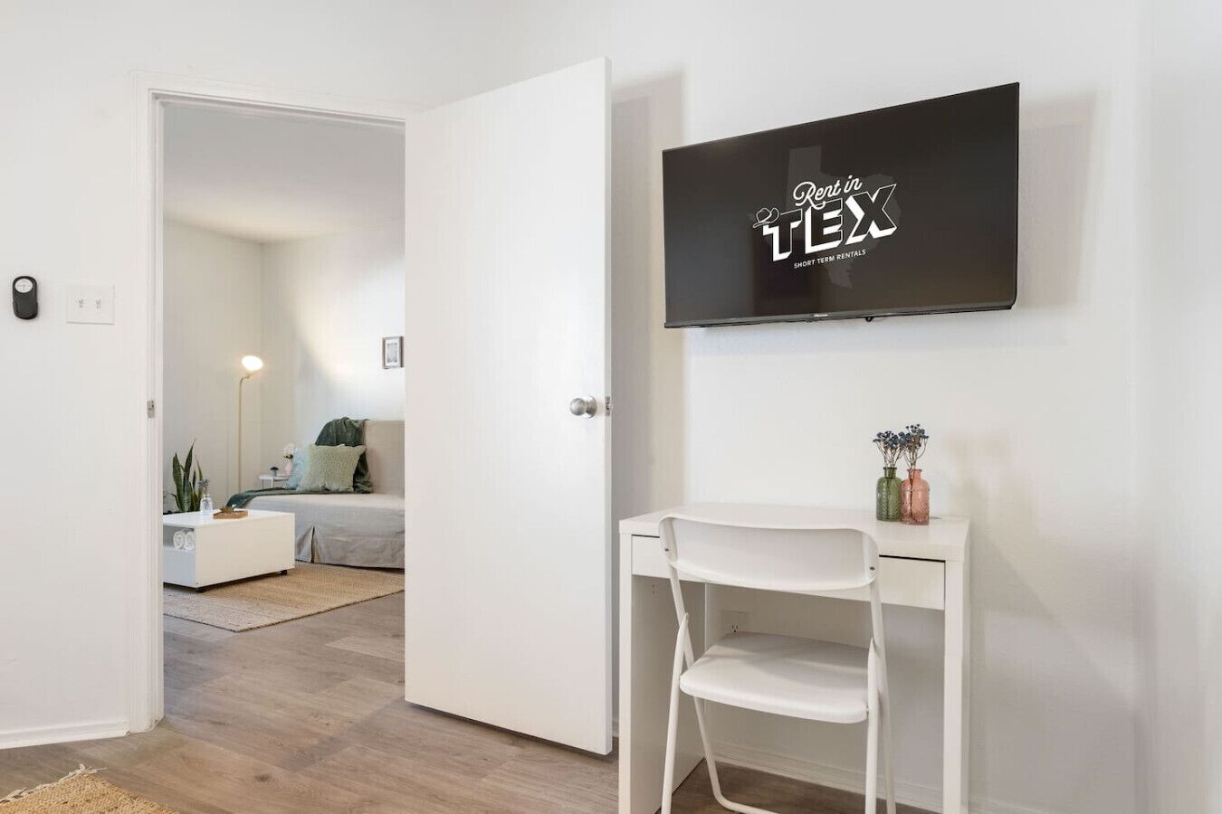 A modern room with a white desk, a chair, a TV mounted on the wall, and a door leading to another room.