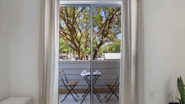 Une pièce dotée d’une porte-fenêtre coulissante donnant sur un espace extérieur aménagé avec deux tables et chaises en bois, un banc blanc et un pot de fleurs.