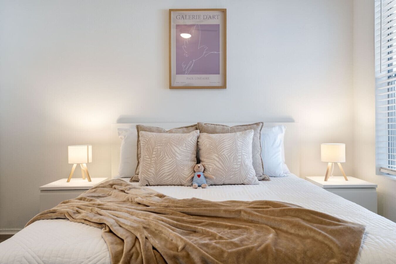 A bedroom with a large bed, two bedside tables, a nightstand, a wall-mounted artwork, and a window with blinds.