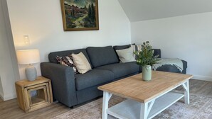 A living room with a grey sofa, a wooden coffee table, a patterned rug, a small side table with a lamp, and a painting on the wall. - Hermine vacation apartment with far-reaching views of nature (Soltau)
