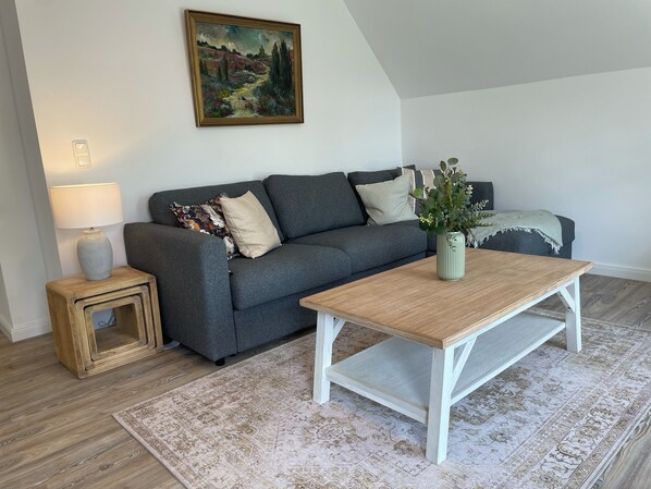 Una sala de estar con un sofá gris, una mesa de centro de madera, un tapiz estampado, una mesita auxiliar con una lámpara y un cuadro en la pared.