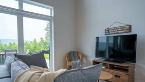 A living room with a large window, a flat-screen TV on a wooden stand, and a cozy seating area with a sofa and armchairs. - Mountain View & Spa | Chalet Le Petit Leo (Saint-Côme)