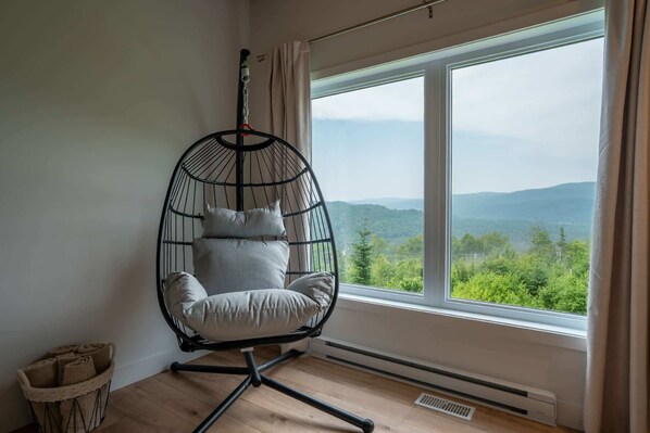 A room with a large window offering a view of green hills, a hanging chair with cushions, and a woven basket on the floor.