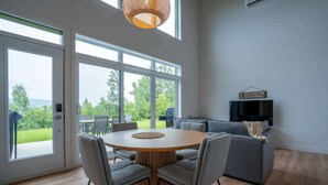 A modern living room with a round wooden table surrounded by chairs, a grey sofa, and a television on the wall. Large windows provide a view of the outdoors. - Mountain View & Spa | Chalet Le Petit Leo (Saint-Côme)