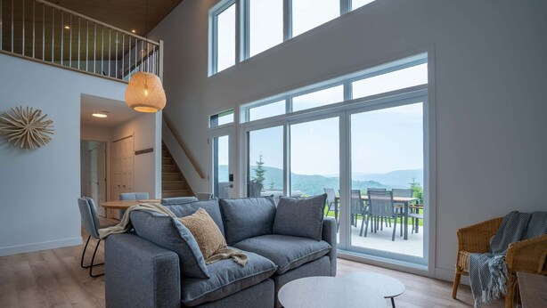 A spacious living room with a grey sectional sofa, a round coffee table, and large windows offering a view of the outdoors. - Mountain View & Spa | Chalet Le Petit Leo (Saint-Côme)