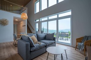 A spacious living room with a grey sectional sofa, a round coffee table, and large windows offering a view of the outdoors.