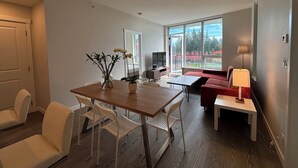 Una sala de estar moderna con comedor, sofá, televisor y un amplio ventanal con vistas a árboles.