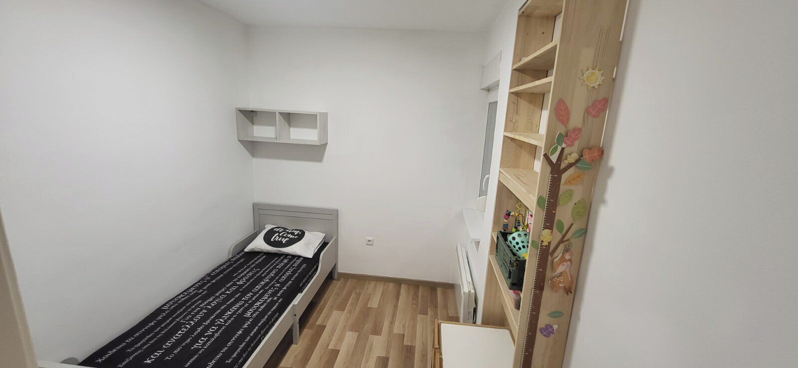 Una habitación pequeña con una cama individual, suelo de madera, pared blanca y una estantería sobre la cama.
