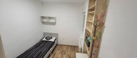 Una habitación pequeña con una cama individual, suelo de madera, pared blanca y una estantería sobre la cama.
