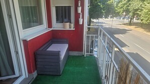Un pequeño balcón con piso de césped artificial verde, un banco de madera y una ventana con persianas.