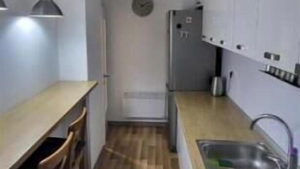 Una cocina estrecha con encimera de madera, un fregadero y un refrigerador. Hay un reloj en la pared y una lámpara colgante sobre la encimera.