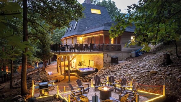 A cozy outdoor seating area with a fire pit, surrounded by Adirondack chairs and a hot tub, set against a backdrop of a house with a balcony and a stone pathway. - Super Luxury Chateau HT/Sauna/Theater/GmRm/4.5Bath (McGaheysville)